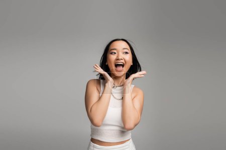 Foto de Excited asian woman in white tank top holding hands near face and screaming isolated on grey - Imagen libre de derechos