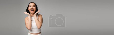 Photo for Astonished asian woman shouting while looking away and holding hands near face isolated on grey, banner - Royalty Free Image