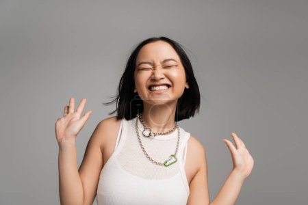 Photo for Excited asian woman in white tank top and metal necklaces showing wow gesture with closed eyes isolated on grey - Royalty Free Image