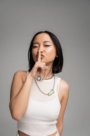brunette asian woman in silver necklaces and white tank top showing hush sign isolated on grey