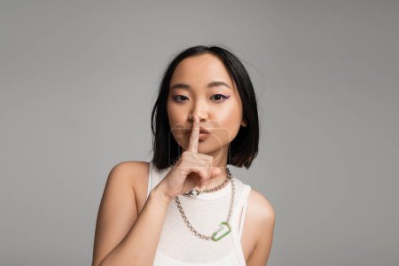 Photo for Young asian woman in white tank top and metal necklaces showing hush sign isolated on grey - Royalty Free Image