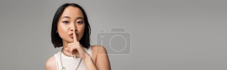 young asian woman with makeup and blue eyeliner showing hush sign isolated on grey, banner