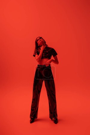 Téléchargez les photos : Full length of stylish woman in latex crop top and trousers posing on red - en image libre de droit