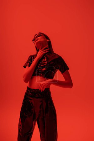 young and stylish woman in latex crop top and trousers touching neck isolated on red