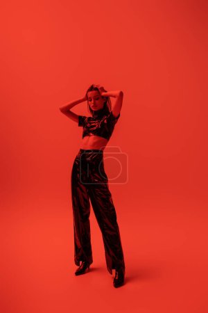 full length of stylish woman in latex crop top and trousers posing with hands on head on red