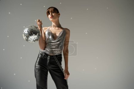 stylish woman in shiny top and leather pants holding disco ball on grey background  Poster 632598534