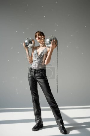 full length of stylish woman in trendy top and leather pants holding shiny disco balls on grey 