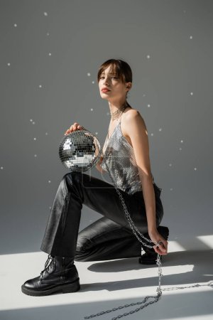 full length of young woman in trendy top and leather trousers holding chain with shiny disco ball on grey 