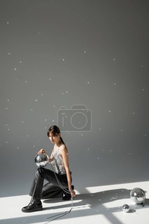 full length of brunette woman in trendy top and leather trousers holding chain with shiny disco ball on grey 