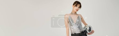 Téléchargez les photos : Brunette young woman in trendy top and leather pants holding retro disco ball isolated on grey, banner - en image libre de droit