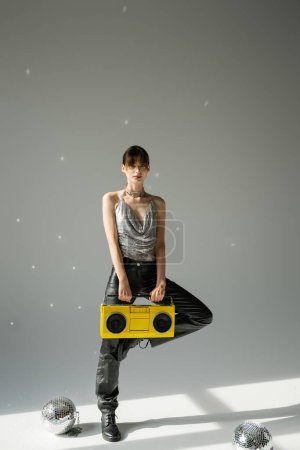 full length of model in trendy top and leather pants holding boombox near disco balls on grey 