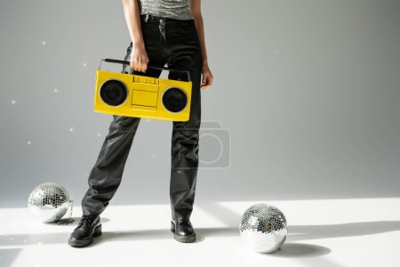 cropped view of woman in black leather pants holding boombox near disco balls on grey 