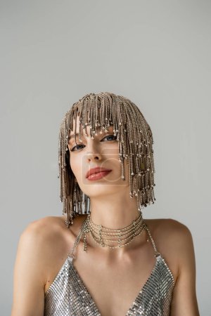 Trendy model in sparkling top and jewelry headwear looking at camera isolated on grey  