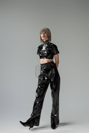 Full length of stylish woman in jewelry headwear and black latex outfit on grey background 