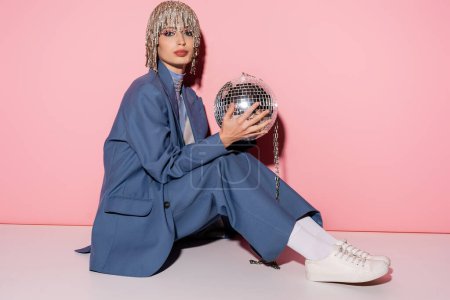 Full length of stylish woman in headwear and suit holding disco ball on pink background 