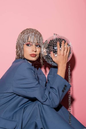 Stylish woman in jewelry headwear and jacket holding disco ball on pink background 