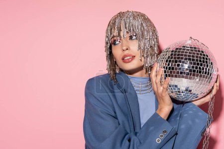 Trendy young woman in jewelry headwear holding disco ball on pink background 