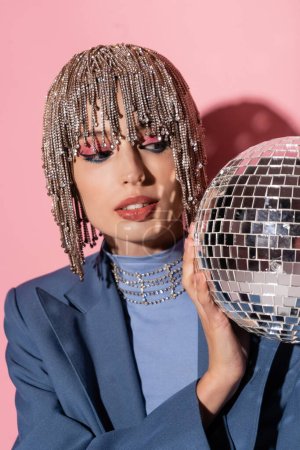 Trendy young woman in jewelry headwear looking at disco ball on pink background 