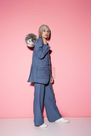 Full length of woman in blue suit and jewelry headwear posing with disco ball on pink 
