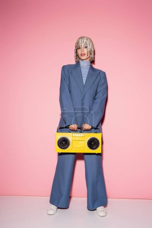 Stylish woman in suit and jewelry headwear holding yellow boombox on pink 