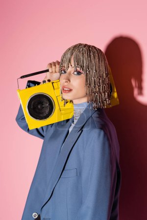 Trendy woman in jacket and jewelry headwear holding boombox on pink background 