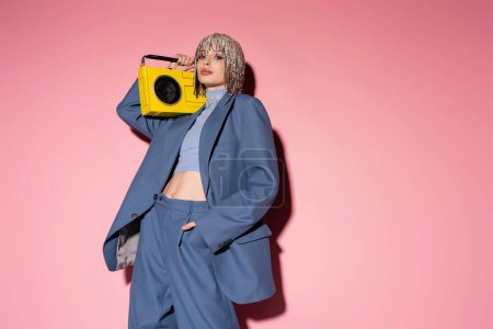 Stylish woman in suit and jewelry headwear holding boombox and posing on pink background 