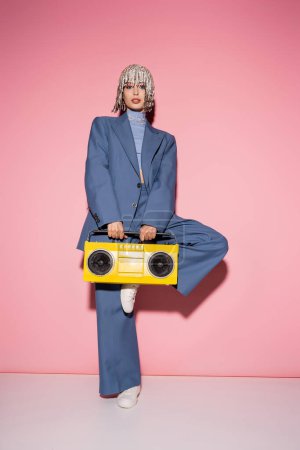 Full length of stylish model in jewelry headwear holding bright boombox on pink background 