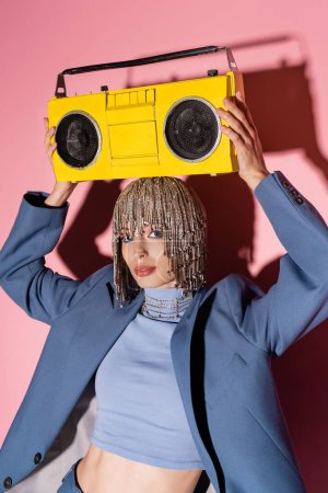 Stylish model in jewelry headwear and jacket holding boombox above head on pink background 