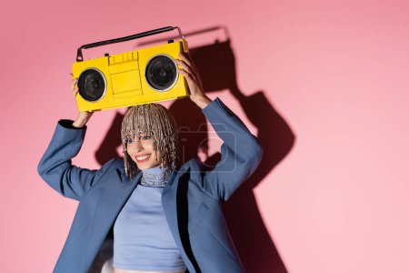 Cheerful young woman in luxury headwear and blazer holding boombox above head on pink background 