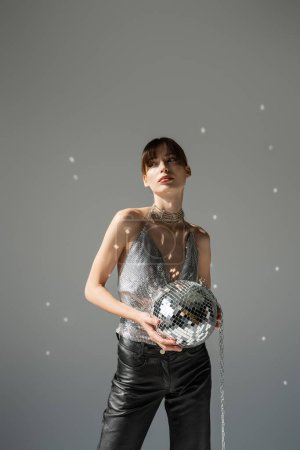 Trendy young woman in shiny top holding disco ball on grey background 