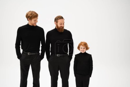 Photo for Redhead bearded men and cheerful boy in black clothes standing with hands in pockets isolated on grey - Royalty Free Image