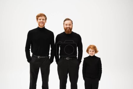 happy bearded men and red haired boy in black clothes posing with hands in pockets isolated on grey