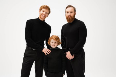 redhead boy standing with hands in pockets near happy dad and grandpa while smiling at camera isolated on grey