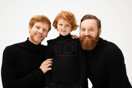red haired and bearded men in black turtlenecks looking at camera near happy kid isolated on grey