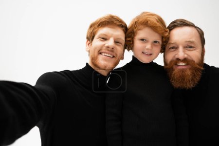cheerful redhead kid looking at camera near bearded father and granddad in black turtlenecks isolated on grey