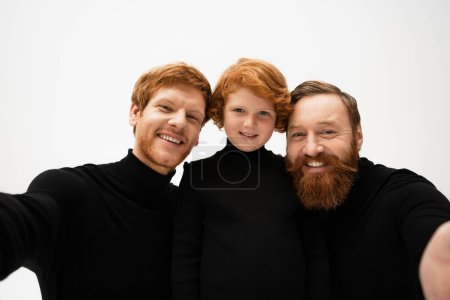 Foto de Joyful bearded men in black pullovers smiling at camera near red haired boy isolated on grey - Imagen libre de derechos
