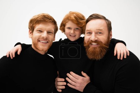Foto de Smiling redhead kid hugging bearded grandpa and dad wearing black turtlenecks isolated on grey - Imagen libre de derechos