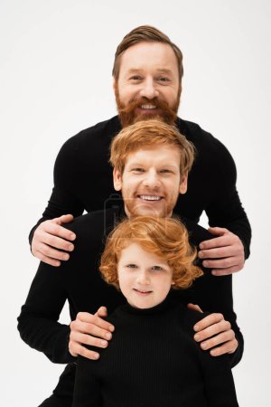 Photo for Happy men in black sweaters smiling at camera near redhead boy while posing isolated on grey - Royalty Free Image