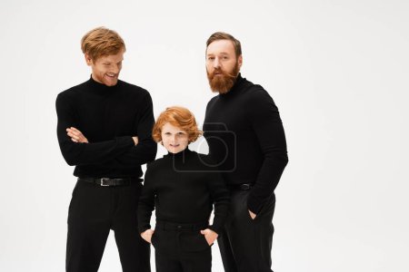 Photo for Bearded man looking at camera near redhead son and grandson posing in black clothes isolated on grey - Royalty Free Image