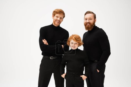joyful redhead kid standing with hands in pockets near bearded dad and grandfather isolated on grey
