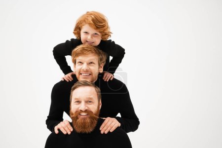 redhead and bearded father and son in black pullovers posing with cheerful kid isolated on grey