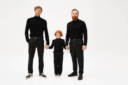 full length of bearded men in black clothes holding hands with joyful redhead boy on light grey background