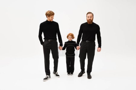 full length of bearded men in black turtlenecks and trousers holding hands with happy red haired kid on light grey background