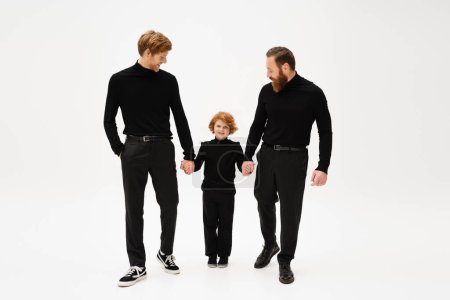 full length of happy redhead kid holding hands with bearded grandfather and dad wearing black clothes on light grey background