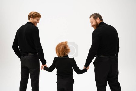 back view of bearded men and red haired boy holding hands and looking at each other isolated on grey