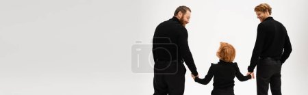Photo for Back view of redhead boy holding hands with smiling father and bearded granddad in black clothes isolated on grey, banner - Royalty Free Image