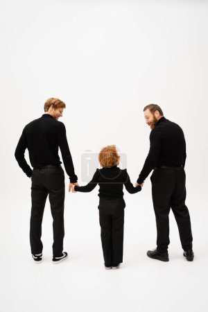 full length of bearded men in black clothes smiling at redhead boy while holding hands on light grey background
