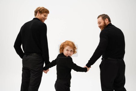 bearded men in black clothes holding hands with joyful redhead boy smiling at camera isolated on grey