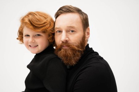 family portrait of bearded man with smiling red haired grandson in black turtlenecks smiling at camera isolated on grey