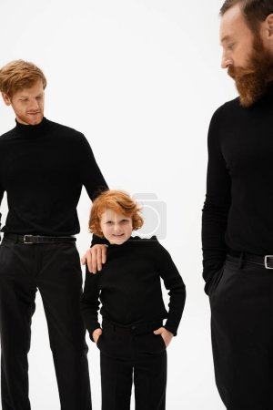 cheerful redhead boy posing with hands in pockets of black trousers near dad and bearded grandpa on blurred foreground isolated on grey